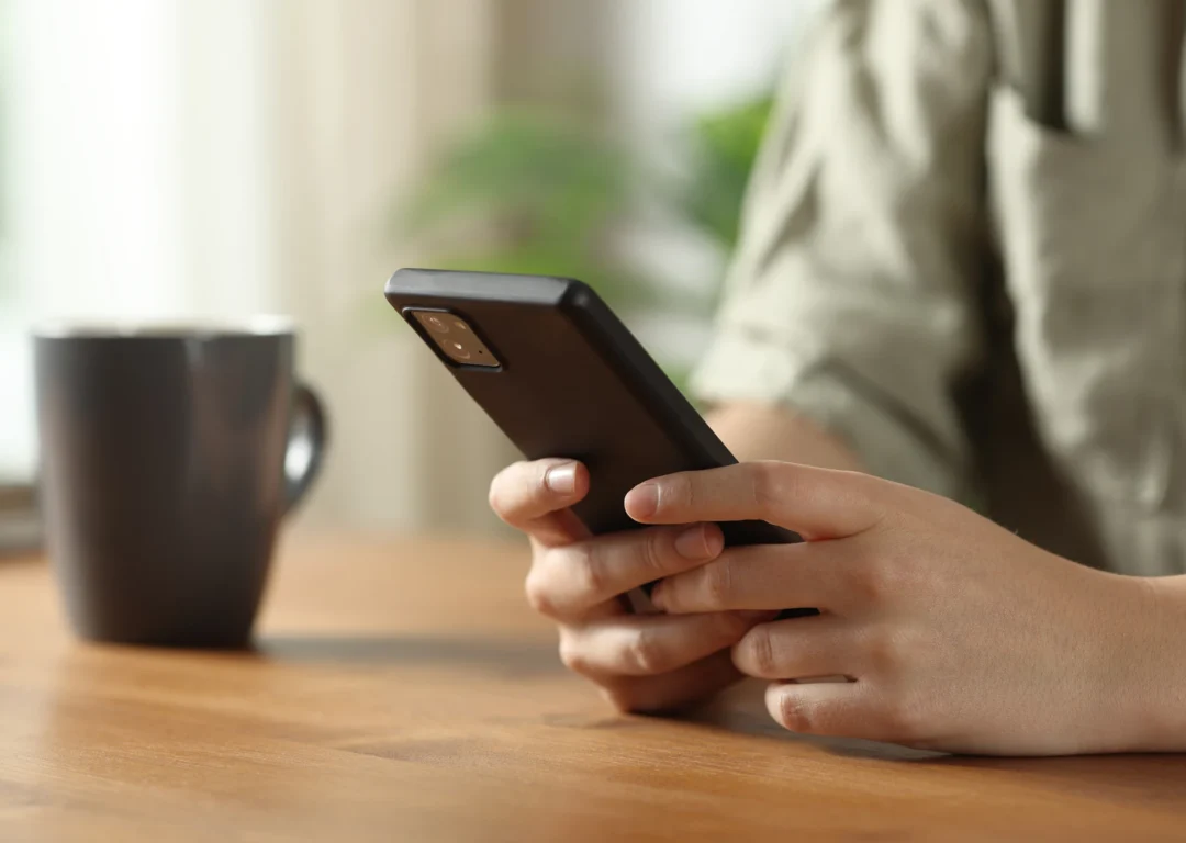 A person sitting at a table, focused on their phone, with a neutral background.