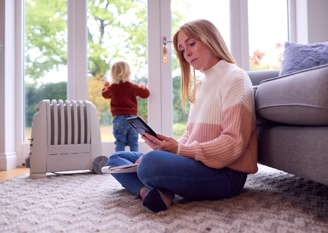 A woman sits on the floor, focused on a tablet screen while her child looks out of the window.