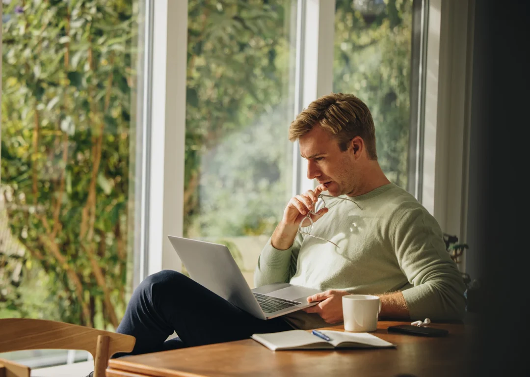 A man sits at a table with a laptop and a cup of coffee, focused on his screen in a home environment.